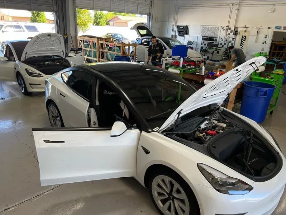White Tesla Model 3 with its doors and frunk open in the Blue Chip Auto Glass shop, with more Teslas and glass crates behind it
