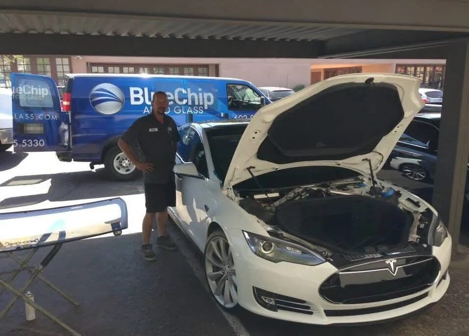 Blue Chip technician beside a white Tesla Model S with its hood open under a carport, with a Blue Chip van behind