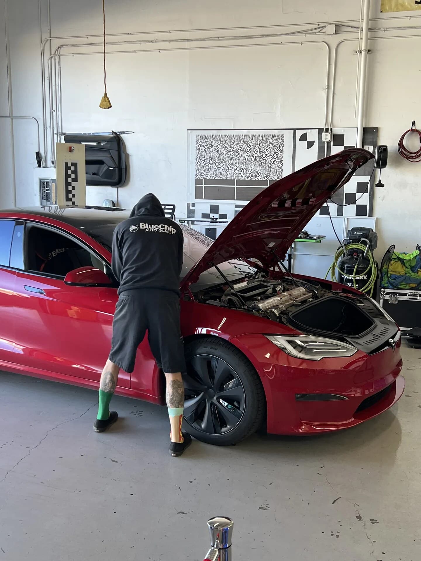 Blue Chip technician working under the open hood of a red Tesla Model S in the shop