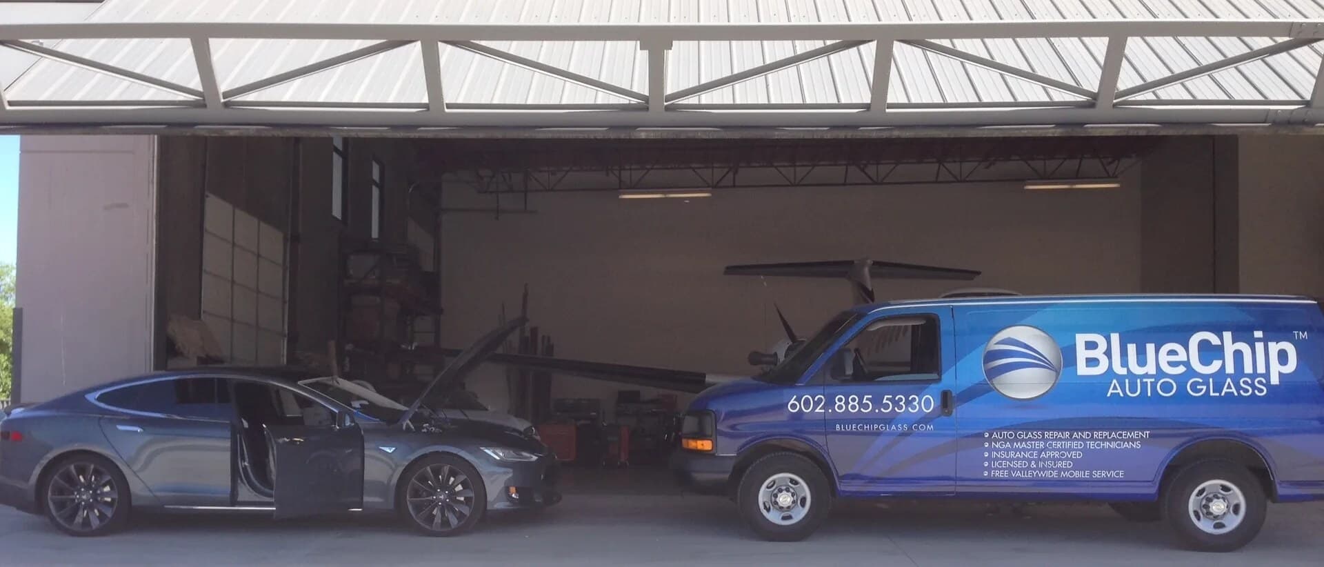 Grey Tesla Model S with its doors and hood open beside a Blue Chip Auto Glass van at an airplane hangar