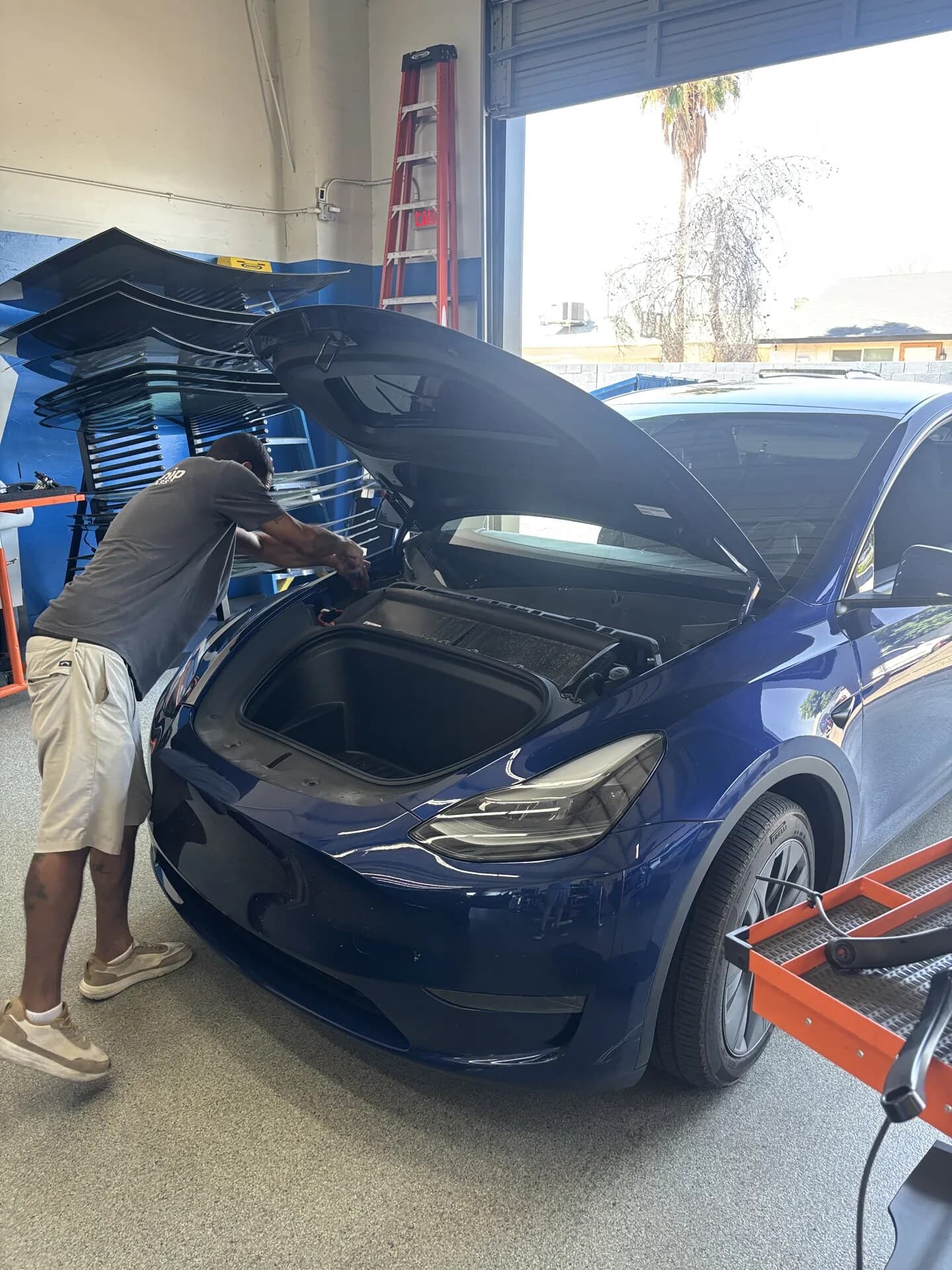 Blue Chip technician working on a blue Tesla Model Y with the frunk open, Tesla windshields racked behind it